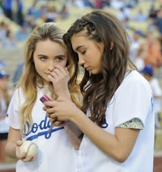 two girls in baseball uniforms looking at their cell phones while standing next to each other