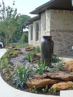 a large vase sitting in the middle of a flower bed next to a building with cars parked nearby