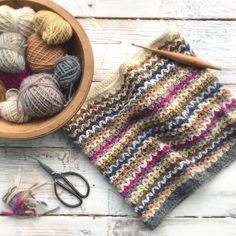 a bowl full of balls of yarn next to a pair of scissors and knitting needles