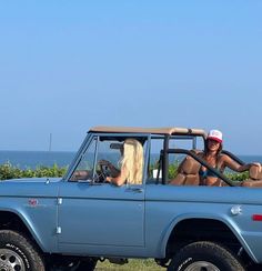 two women sitting in the drivers seat of a blue pickup truck with an ocean view behind them