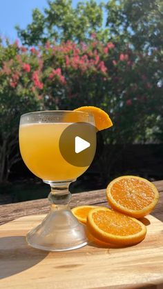 an orange juice in a glass next to sliced oranges on a wooden table with trees in the background