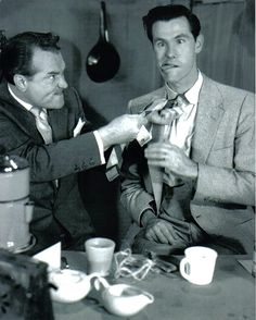 two men are sitting at a table and one is tying the tie on another man's neck