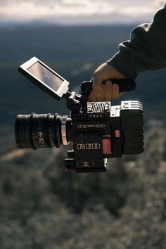 a person holding a camera with a cell phone attached to it's handle, in front of some mountains