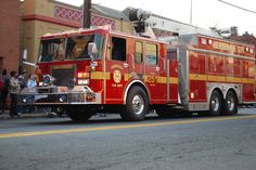 a red fire truck driving down a street next to a tall brick building with people standing around it