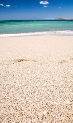 footprints in the sand on a beach with clear blue water and white clouds behind it