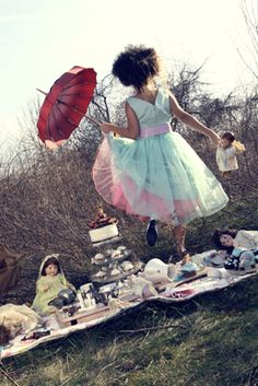 a woman in a dress jumping over a picnic table with an umbrella and other items on it