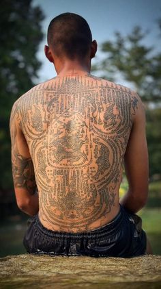 a man with tattoos on his back sitting in front of a body of water and trees