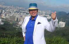 a man in a white suit and blue shirt standing on top of a hill with his hand up