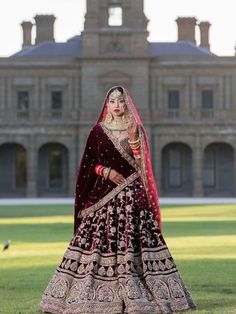 a woman in a red and gold bridal outfit standing on grass with a large building in the background