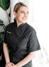 a woman standing in front of a window wearing a black chef's coat and smiling at the camera