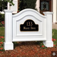 a white and black sign in front of a house with red flowers around the yard