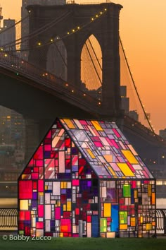 a house made out of stained glass sits in front of the brooklyn bridge at sunset