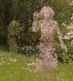 a woman standing in the middle of a field with flowers around her and trees behind her