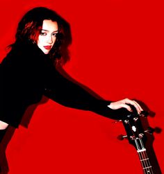 a woman holding a guitar in her right hand and posing for the camera on a red background