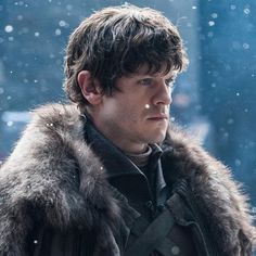 the young man is wearing a fur coat and looking off into the distance with snow falling on his face