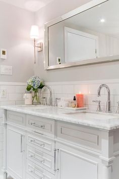 a white bathroom with two sinks and a large mirror above the sink is lit by a candle