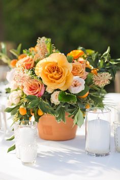 an arrangement of flowers in a pot on a white table cloth with candles and glasses