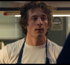 a young man with curly hair wearing an apron and looking at the camera while standing in front of him