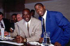 two men in suits sitting at a table with drinks and papers on the table, smiling