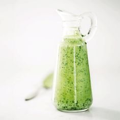 a green liquid in a glass pitcher with a spoon next to it on a white surface