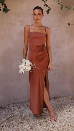 a woman standing in front of a wall wearing a brown dress and holding a bouquet