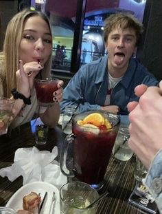 three people sitting at a table with drinks and food in front of them, while one person holds his mouth open