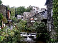 a river running through a lush green forest covered hillside next to tall brick buildings with signs on them