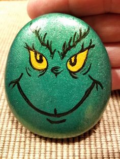 a hand holding a green rock with yellow eyes and grin face painted on it's side