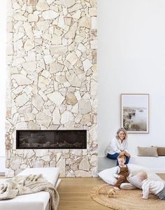 two people are sitting on the floor in front of a fireplace with a stone wall behind them