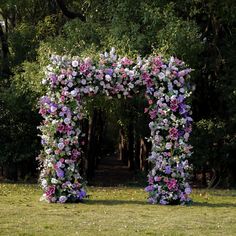 an arch made out of pink and purple flowers in the grass with trees in the background