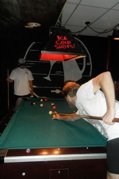 a man leaning over to hit a ball on a pool table with two other men in the background