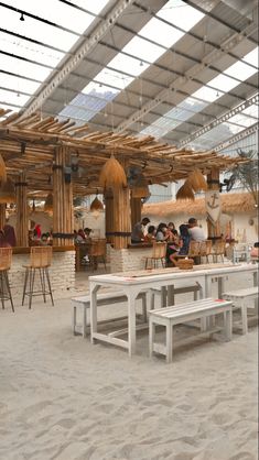 people are sitting at tables in the sand under an open roof area with straw umbrellas