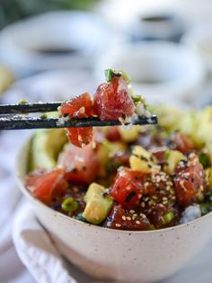 someone is holding chopsticks over a bowl of food that includes tomatoes, avocado and sesame seeds