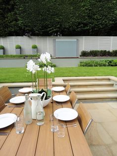 a wooden table with white plates and vases on it in front of a garden