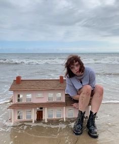 a woman sitting on top of a doll house in the sand at the beach near the ocean