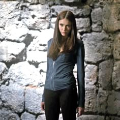 a woman standing in front of a stone wall with her hands on her hips and looking at the camera