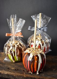 two caramel apples wrapped in cellophane and sitting on a wooden table with candy sticks sticking out of them