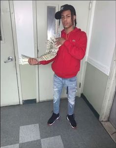 a man in a red shirt and black hat is holding up some money while standing in front of a door