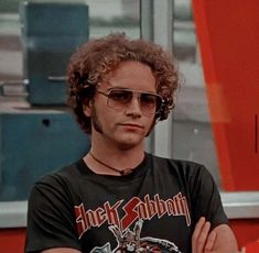 a man with curly hair and sunglasses standing in front of an orange wall holding his arms crossed