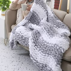 a woman sitting on a couch holding a coffee cup in her hand while she is wrapped up in a gray and white crocheted blanket