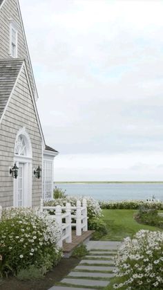 a white picket fence next to a house with flowers in the foreground and water in the background