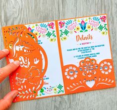 a person is holding up an orange and white wedding card with flowers on the front