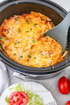 an open crock pot filled with cheesy casserole next to tomatoes and lettuce