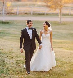 a bride and groom holding hands walking through the grass in front of a fenced area