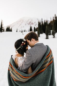 a bride and groom wrapped in a blanket on top of a snow covered hill with trees