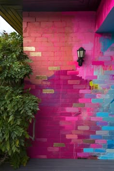 a pink and blue painted brick wall next to a green bush with a lamp on it