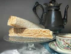 two sandwiches on a glass plate next to a cup and saucer with a teapot in the background