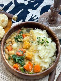 a wooden bowl filled with stew next to biscuits and bread on a tablecloth covered place mat