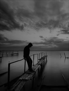 a man standing on a wooden dock in the middle of water at dusk with dark clouds overhead