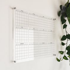 a wall mounted calendar on a white wall next to a potted plant with green leaves